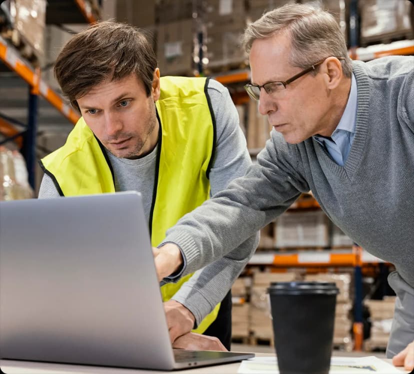 Two men checking a laptop in a warehouse, one wearing a high-visibility vest